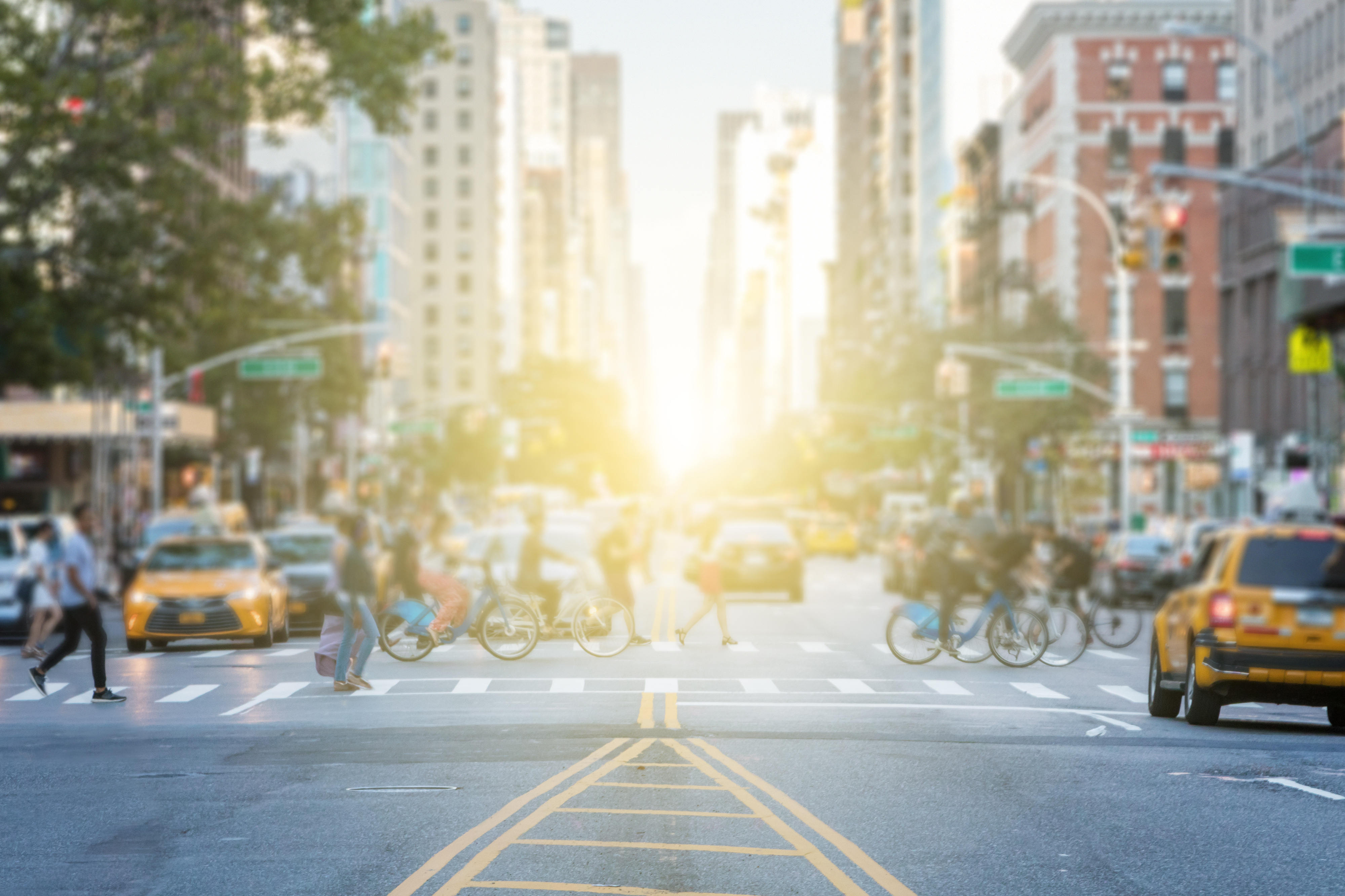 busy crosswalk with pedestrians and bikers crossing in front of traffic, bright sun between streets lined with tall city buildings. Image Credit: AdobeStock_172277527
